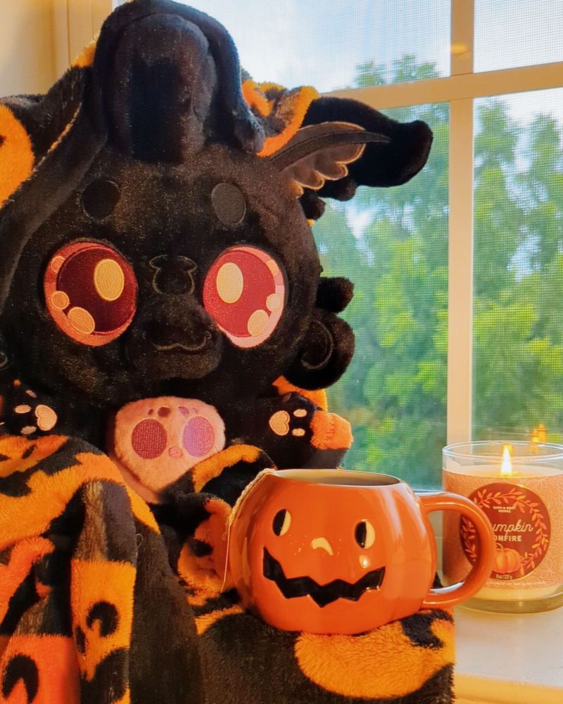 Mothman plush sitting in jack-o'-lantern Halloween blanket, with a mug of steaming tea in and jack-o'-lantern mug - accompanied by a pumpkin bonfire scented candle.