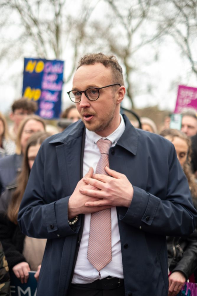 Adam Dance MP speaking with emotion at a public event, hands on his chest, surrounded by a crowd. Protest signs can be seen in the background with messages including “No child left behind.”