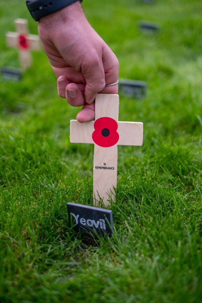 Adam Dance's hand plants a remembrance cross with a red poppy in the ground, next to a marker saying 'Yeovil'. The poppy has 'In Remembrance' on it in black writing.
