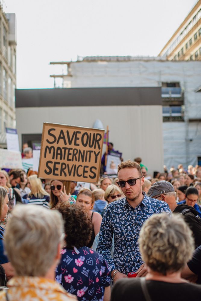 Adam Dance MP speaks with members of the public during a protest. A large crowd surrounds him, with one person holding a sign that reads "SAVE OUR MATERNITY SERVICES." The event takes place in front of a town hospital building under renovation.