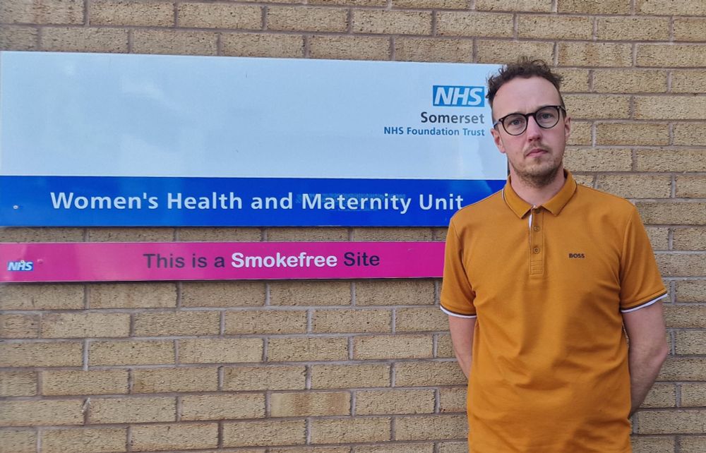 Adam Dance MP stands in front of a sign for the Women’s Health and Maternity Unit at Yeovil District Hospital. The sign also states the area is a smoke-free site. He is wearing a mustard-coloured polo shirt and looking serious.