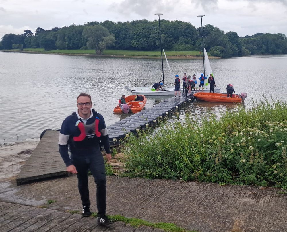 Adam Dance MP walks up a slipway smiling, wearing a life jacket and sailing gear. In the background, a group of people in helmets and vests are preparing to sail small boats on a large lake surrounded by trees.