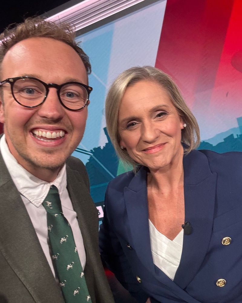 Adam Dance MP takes a smiling selfie with journalist Amanda Parr in a brightly lit studio. Both are dressed smartly, with Amanda wearing a navy blazer and Adam in a green tie with a white shirt and jacket. A red and blue graphic backdrop is behind them.