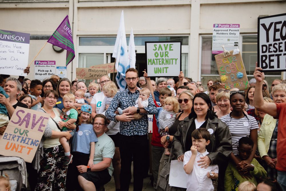 Adam Dance MP stands holding a baby among a large crowd of parents, children, and campaigners outside a hospital building. The group holds handmade signs reading “Save Our Maternity Unit,” “Save Our Birthing Unit,” and “Yeovil Deserves Better,” showing strong community opposition to the closure of Yeovil’s maternity services.