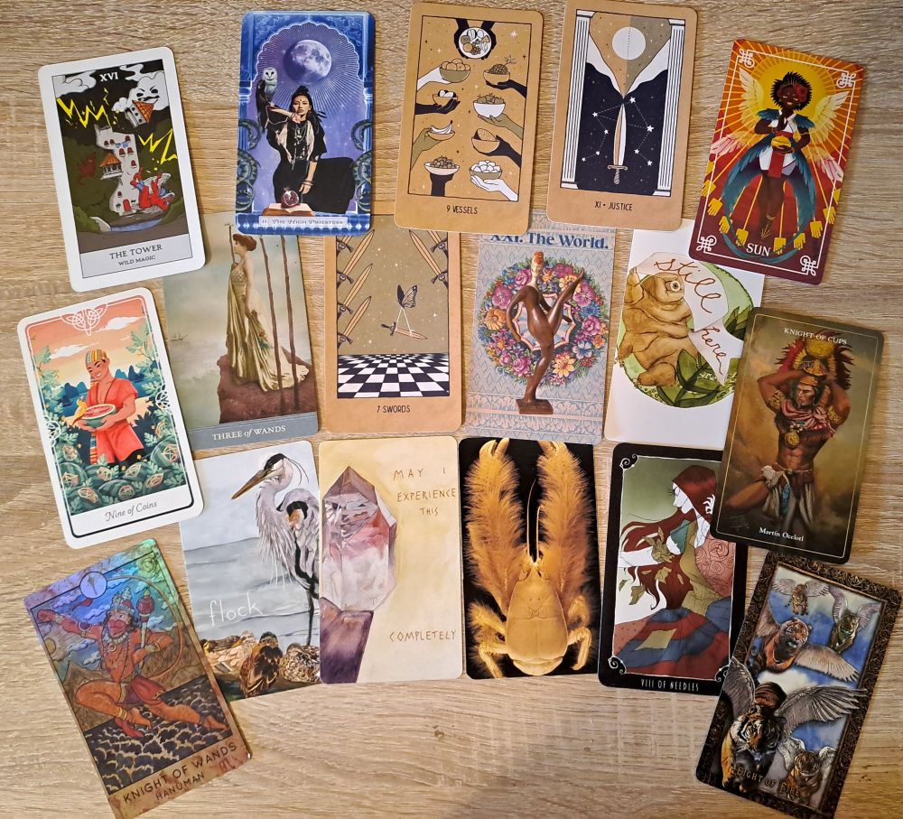 Photo of several tarot cards from different decks spread out on a table. They all feature different symbols and art styles, for example, one has Sally from Nightmare Before Christmas as 8 of Swords, and another is just a photo of a yeti crab.