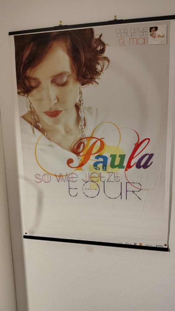 Ein farbenfrohes Poster mit der Aufschrift „Paula so wie jetzt TOUR“. In der oberen rechten Ecke steht kleinerer Text mit „neue im mai“ und „Paula“. Es scheint eine Werbung für eine Tour von Paula zu sein.