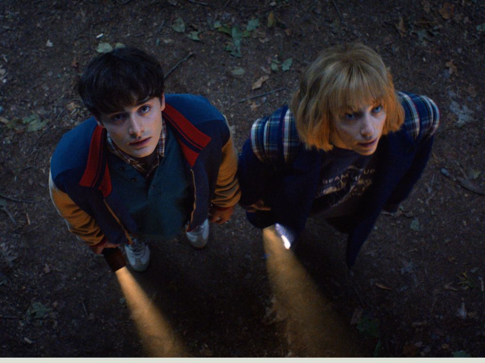 Will and Robin stand in a dirt tunnel, holding flashlights and looking up at Lucas, who is out of frame. 