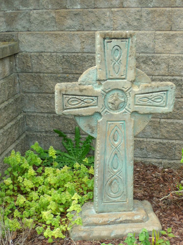 A weathered Celtic cross stands in a garden, surrounded by lush greenery and set against a rustic stone wall. The cross features intricate carvings and a verdigris patina, giving it a sense of age and character. The garden is filled with vibrant plants and flowers, adding a pop of color to the scene. In the background, a stone wall provides a natural backdrop, while the cross remains the focal point of the image.