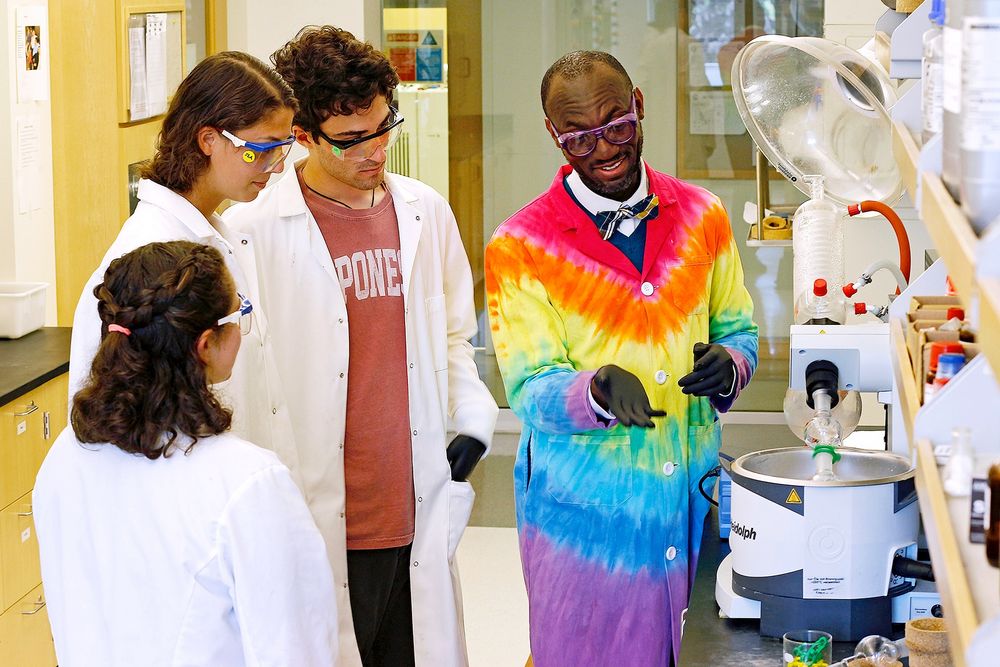 Dr. Andre Isaacs and is wearing a rainbow tie dye lab coat. He appears to be showing a group of three students something on a microscope. The students are wearing white lab coats.