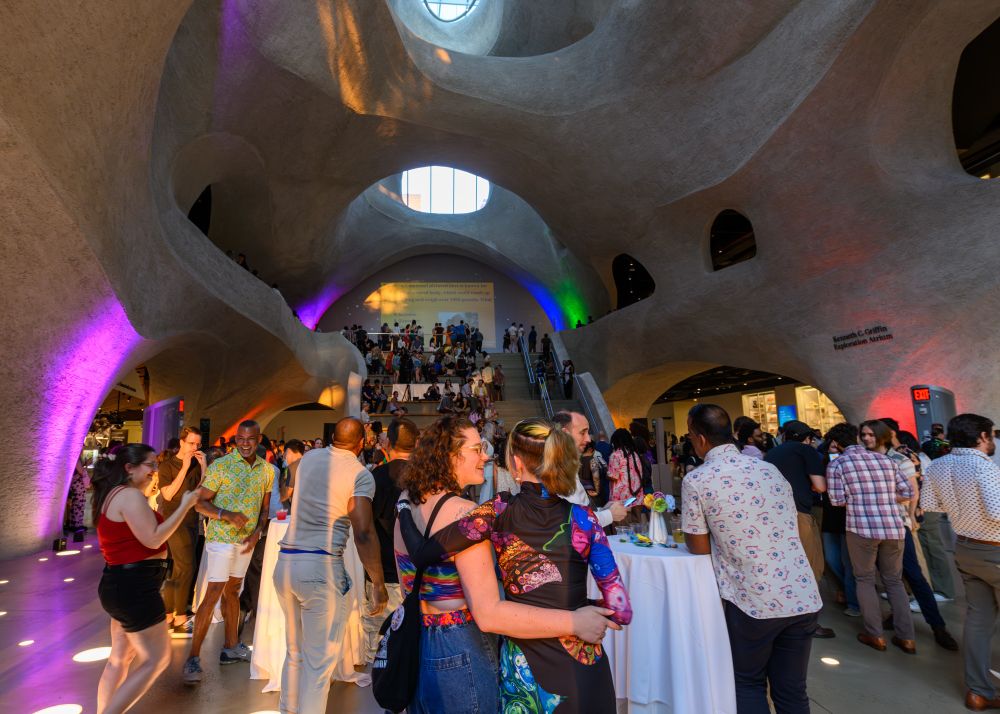 Wide shot of the Griffin Exploration Atrium inside of the Gilder Center. It’s colorfully lit and there’s a crowd of people mingling and dancing on the floor. 
