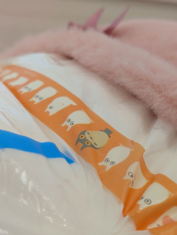 Close up of a plain white ABDL diaper's landing zone. An orange tape featuring Chibi-Totoros in various poses stretches across the front. Out of focus appears to be some fluffy pink pants that have been pulled down to expose this kiddos diaper.