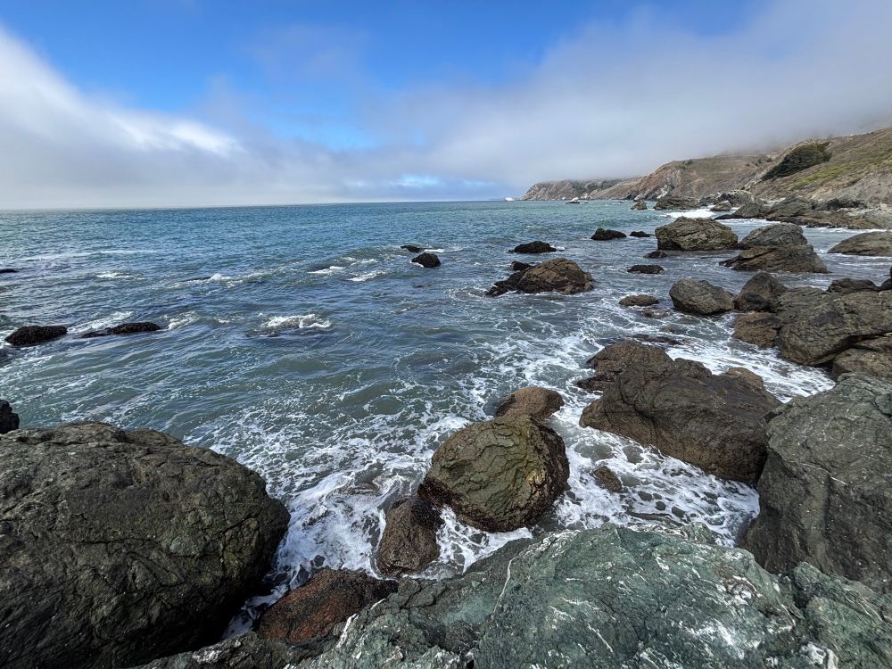 Ocean waves hitting rocks on a rocky coast. Fog above with a little bit of blue sky.