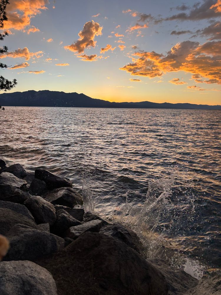 Sunset over mountains in background. Nearby are waves of Lake Tahoe hitting rocks. 