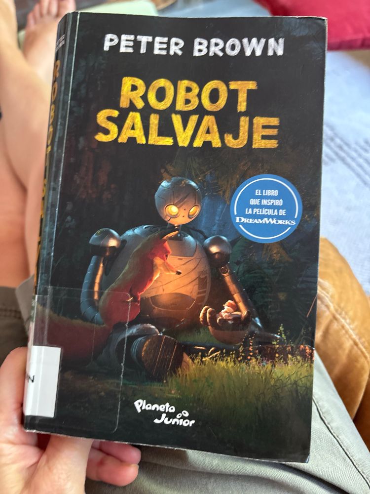 Cover of Robot Salvaje. Illustration is from the movie and shows a large robot holding a tiny bird in one hand. A fox is looking at the bird. 