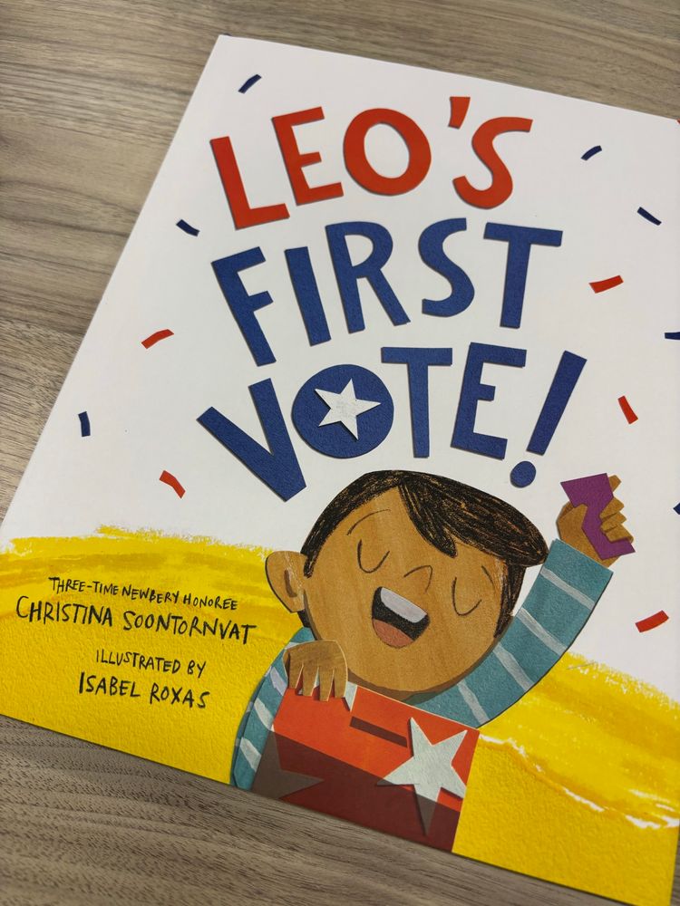 Cover of book Leo’s First Vote. Young brown skinned child holding a ballot up in one hand and a ballot box in the other.
