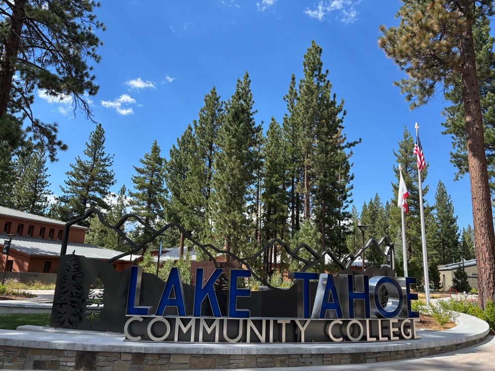 Sign for Lake Tahoe Community College with pine trees and building behind it. Blue sky with a few wispy clouds.