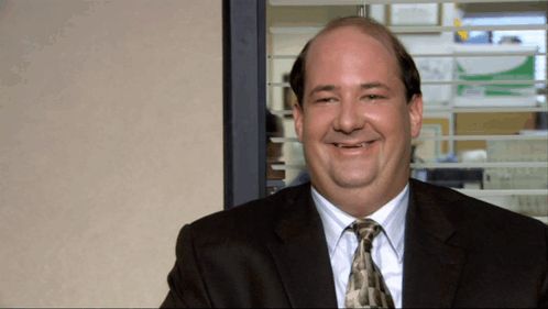 a bald man in a suit and tie smiles in front of a window