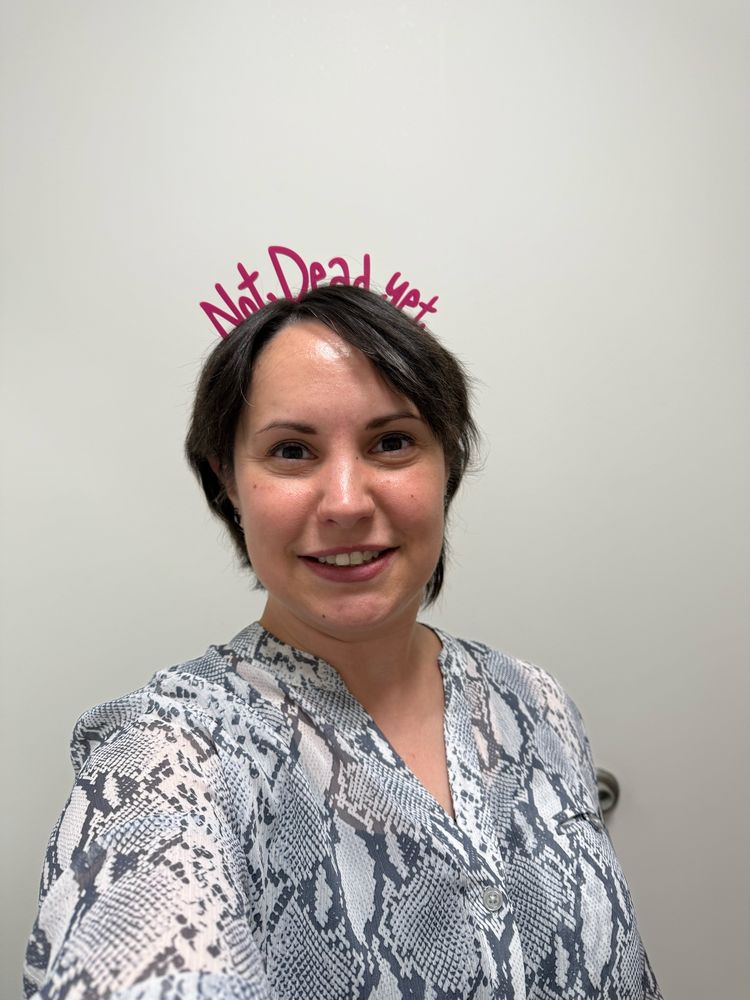Mid-30s woman with short brown hair facing the camera smiling. She is wearing a gray & white snake skin print blouse and a pink headband that says “Not Dead Yet” 