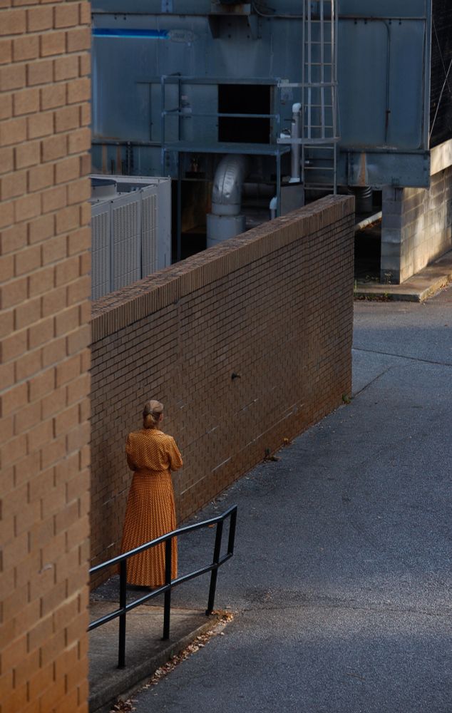 A photograph of a woman in an orange dress taking a smoke break in an orange-brick loading bay