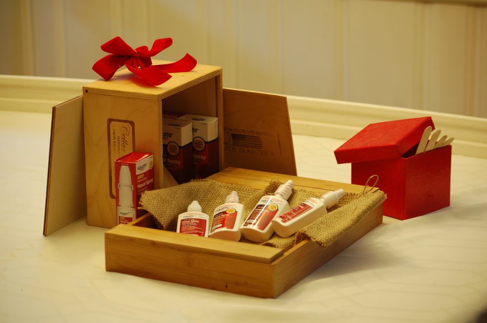 A still life photograph: bottles of nasal decongestant spray are arranged in a wooden box in a manner that somewhat evokes novelty wine bottles, dominant reds and cream colors
