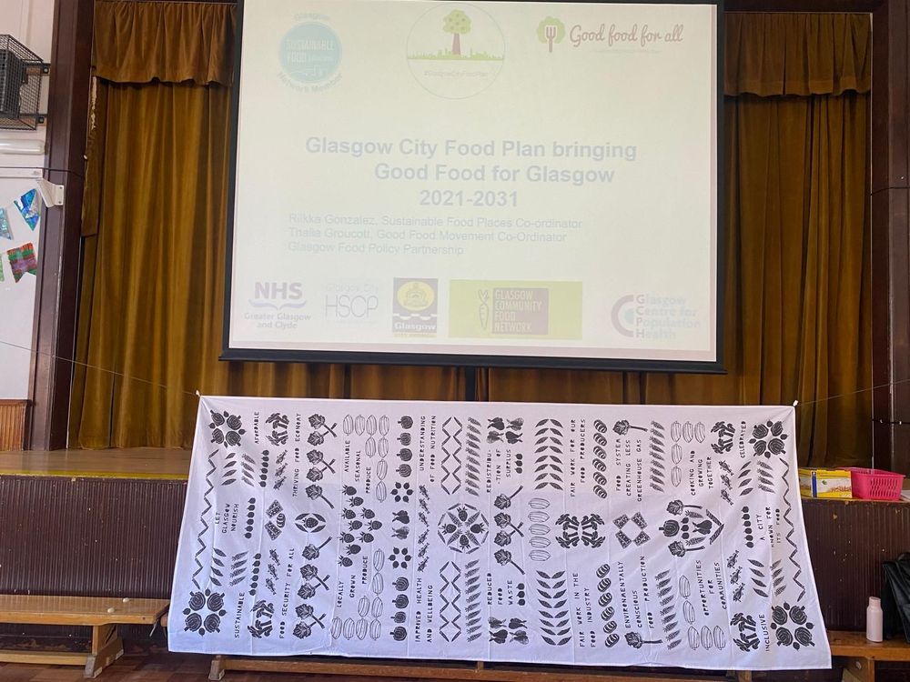 Slide with the Glasgow Food Policy Partnership logo and those of some of our partners - the NHS, HSCP, GCC, GCFN, and GCPH. Text on the slide reads 'Glasgow City Food Plan bringing Good Food for Glasgow 2021-2031. A collaborative tablecloth facilitated by Hannah Brackston is hung across the stage below the projected slide.