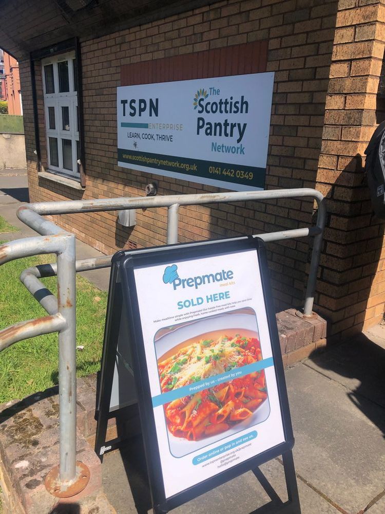 A-board sign with a photo of a meal and text reading 'prepmate sold here' in front of a brick building with a sign for 'TSPN enterprise' and 'The Scottish Pantry Network'
