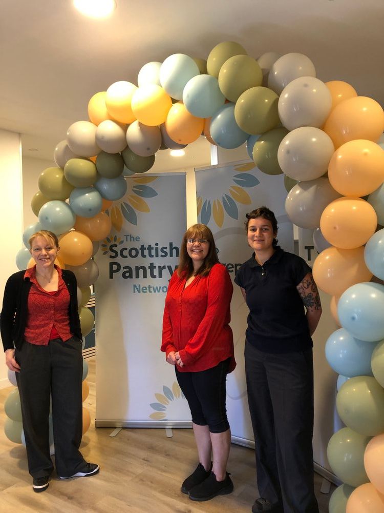 Three people standing under a balloon arch and next to a roll banner which reads 'The Scottish Pantry Network'. They are all smiling and looking at the camera.