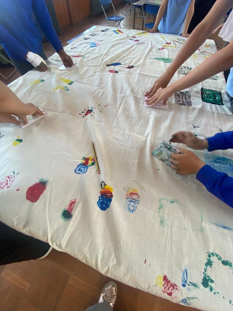 Handprinted food designs including fruit and words across a tablecloth. There are hands reaching into frame which are adding more designs. 