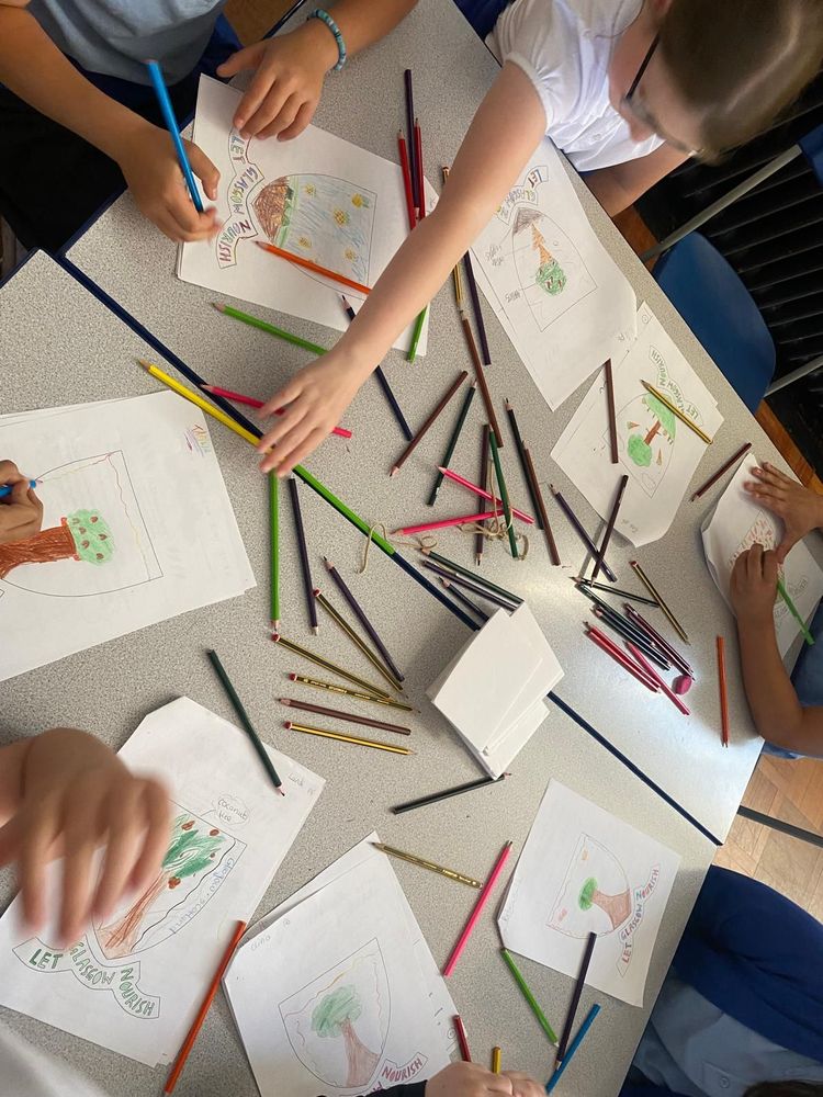 Picture from above of a table with pupils sitting around it, with coloured pencils and Glasgow crest worksheets in front of them. One is reaching across the table for a pencil and the others are out of frame apart from their hands.
