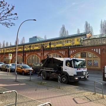 Street scene in Berlin - the yellow U-Bahn train in background on an elevated track over a brick building. Trucks and cars in foreground.

