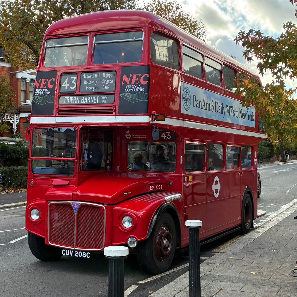 A red "route master" bus showing route 43, on a leafy London street with vintage (1980s?) ads on it for "NEC" computers and "Pan Am".