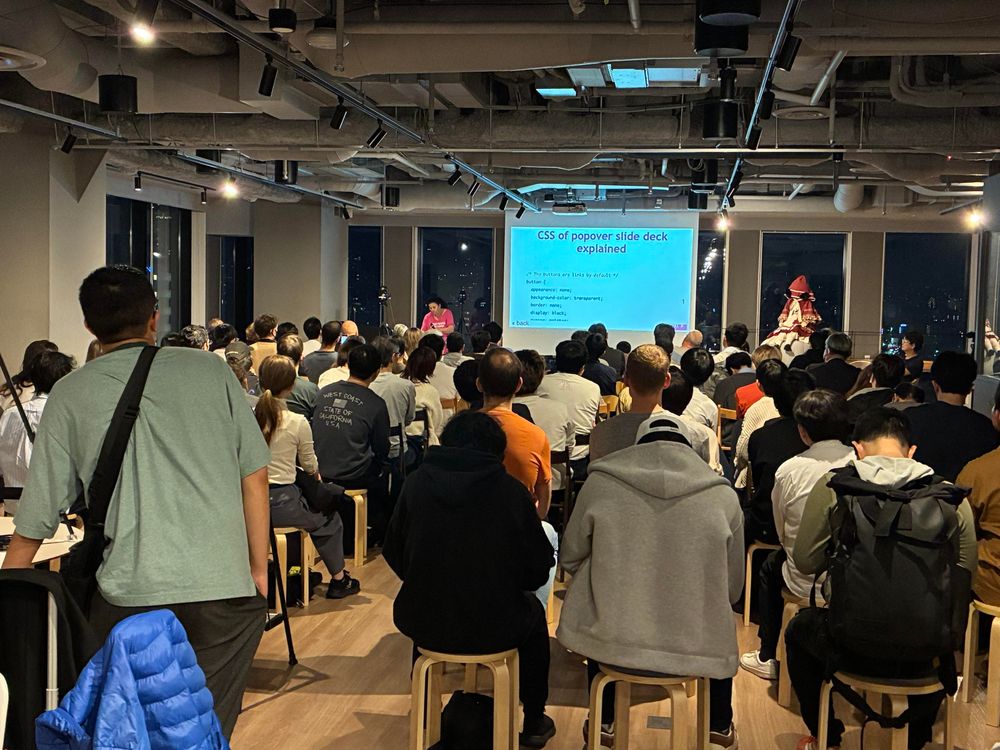 A crowd of people sits in an indoor space, attentively watching a presentation. The presenter, standing on a stage, is visible on the right, while a slide displayed on a screen outlines topics related to CSS and popover slide decks.