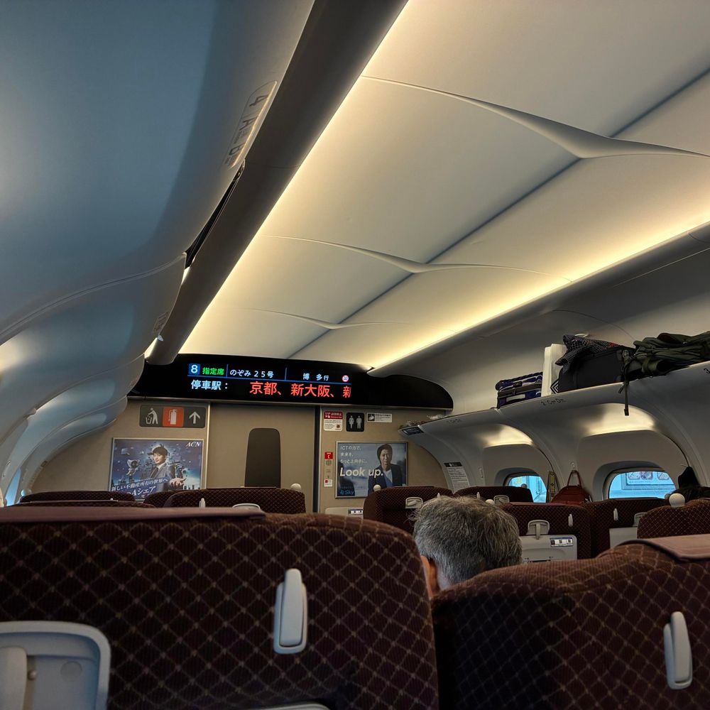 Interior of a train with plush, maroon seats and overhead luggage compartments. A digital display shows route information in Japanese, indicating stops in Kyoto and Osaka. Soft lighting is evident in the ceiling design.