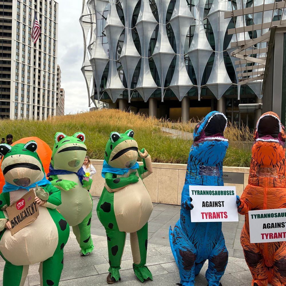 A group of individuals dressed in inflatable dinosaur and frog costumes stands outside a modern building (the US embassy in London). They hold signs that read "TYRANNOSAURUSES AGAINST TYRANTS" and one figure displays a "STOP the broligarchy" sign.
