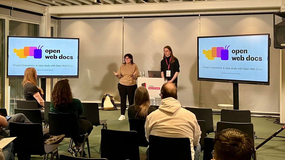 Jory and Florian stand on stage behind a podium (marked "FOSS backstage") with monitors on both sides of them reading "open web docs" and showing the first 2 rows of seating with some attendees watching.