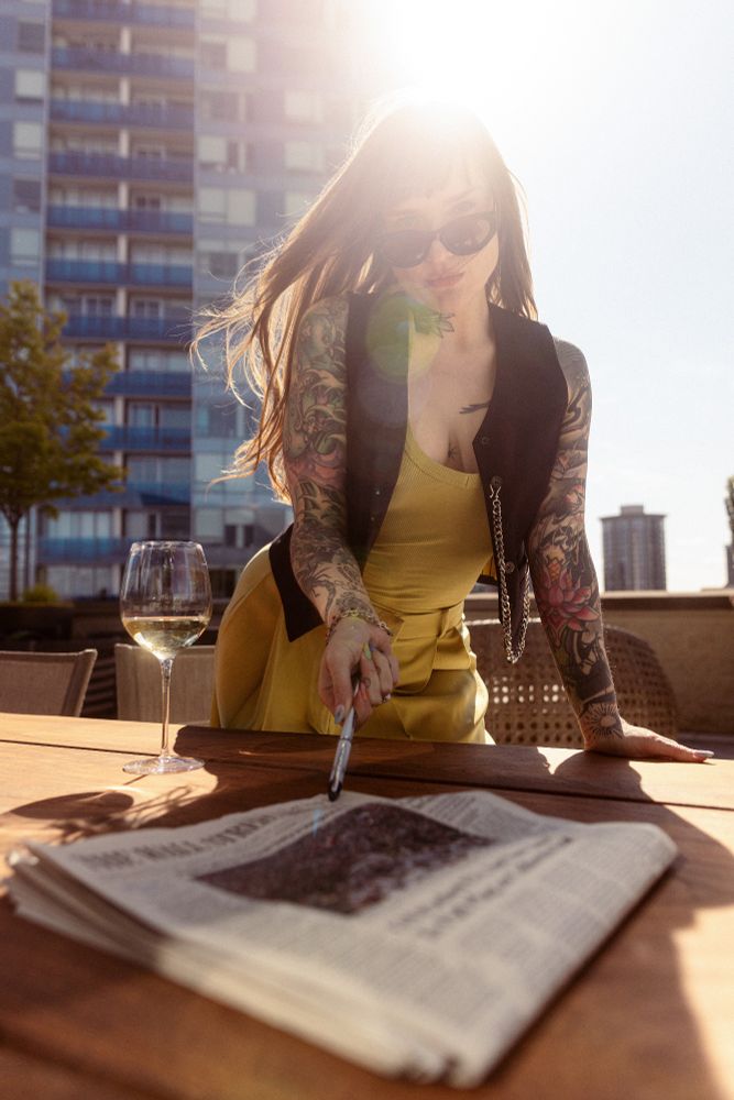Vaughn is on a patio in a city while wearing green pants and a black vest with a silver wallet chain, sunglasses and a green shirt. 
There is a newspaper in the foreground and Vaughn is using a pen to point at the name of the newspaper. 
A glass of white wine is placed off to the side. 