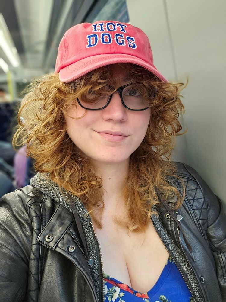 A selfie of me in a blue top, jacket and hat that says hot dogs
