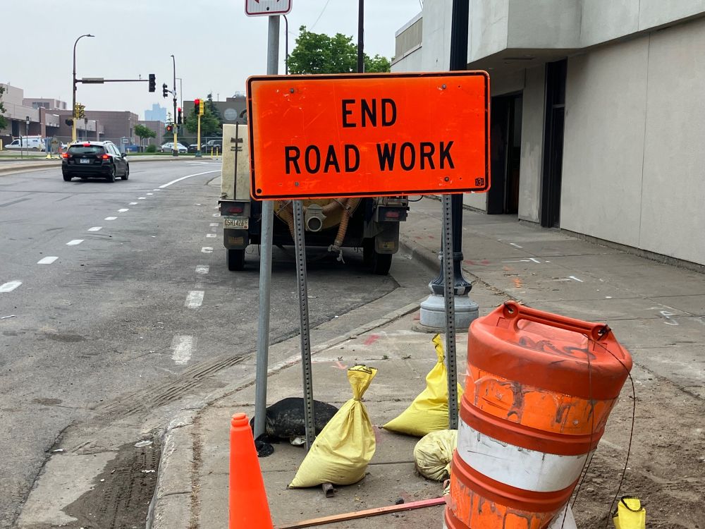 An orange road sign stating “End Road Work” on a south Minneapolis sidewalk with car, road and building in background.