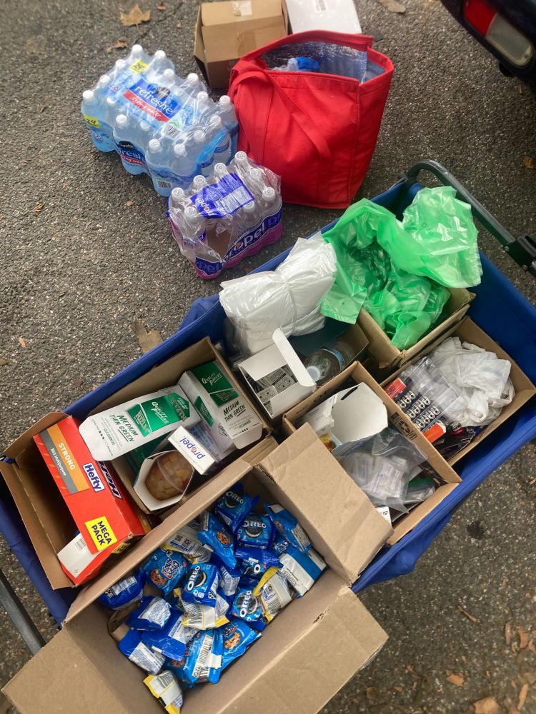 top view of a blue cart with supplies including cookies, rolling papers, trash bags, fruit cups, and more! To the left of the cart are several packages of bottled water and sports drinks