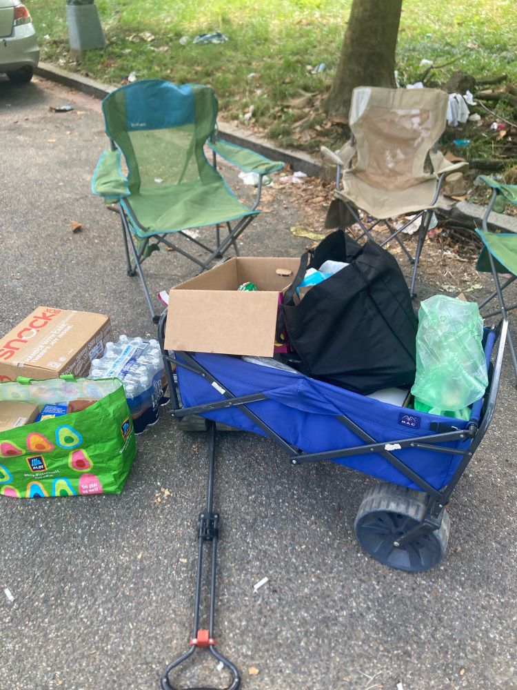 Picture of a blue cart full of supplies sitting next to three camp chair. On the ground next to the cart are several bags of supplies and a package of bottled water.