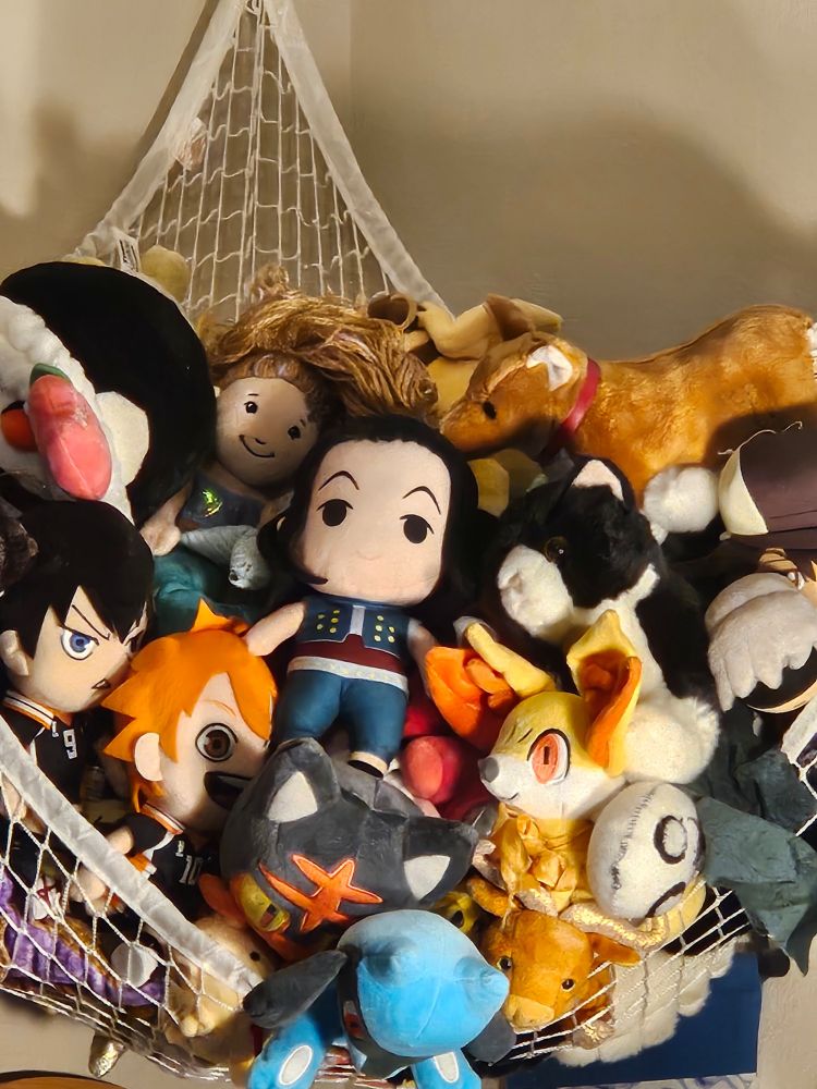 hammock attached to the wall full of various plushies. shown: a penguin in a cute headband, kageyama and hinata from haikyuu, litten, riolu, fennekin, a small purple dragon, an ox, a handmade emil from nier, 9s, a black and white cat, ein from cowboy bebop, a mermaid, kotetsu from tiger and bunny (barnaby is just off camera), illumi from hunterxhunter, and several others too obscured to tell what they are.