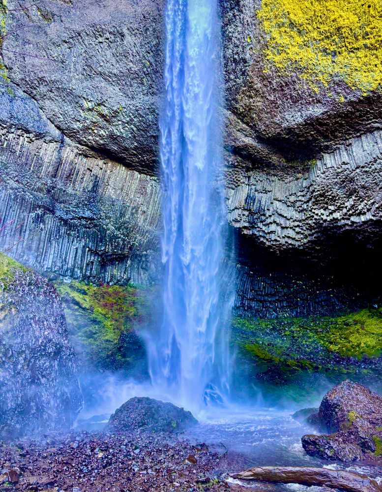 Standing at the bottom of a waterfall with water spraying up as it hits the rocks and a pool of water. The waterfall is bluish white and is surrounded by gray and black basalt rock. There is yellow and green lichen growing on the bottom of the basalt rock here and there, and an especially large yellow patch at the upper right corner.