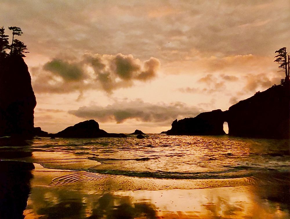 Standing a beach at low tide looking out to the ocean at sunset. There are silhouetted rocks in the ocean to the left, right and in front. A small wave is gently rolling in. And the sky is a muted orange with some puffy clouds which are reflecting orange off the water.