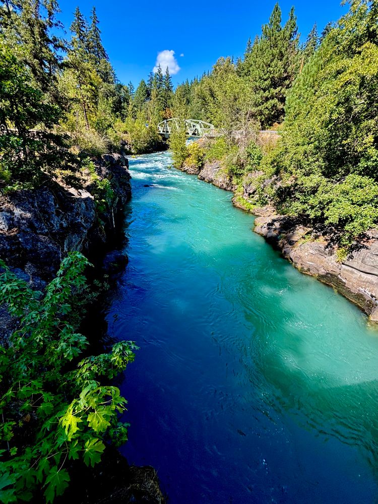 A river with stunning turquoise-colored water runs through the photo with a steel arch bridge at the top end. The river is flanked by rocky cliffs on both sides and evergreen trees are growing on top of the rocks. Bright blue sky with one fluffy puff of a white cloud can be seen above the river and evergreen trees.