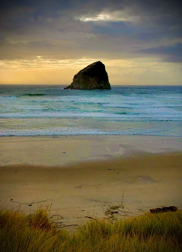 A view of a huge haystack-shaped monolith rock in the Pacific Ocean in the center of the image. There are dark storm clouds overhead at the top with a strip of sunset sky underneath surrounding the rock. The ocean it sits in is an aqua green with gentle white-crested waves rolling onto the sandy beach of tans and light browns. In the foreground are beach grasses of greens, rusts, and golds.