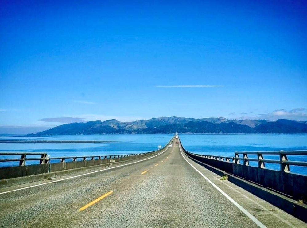 View as I’m driving on a concrete bridge across a very wide blue river. At the other end of the very long seemingly endless bridge are dark blue mountains. The sky is blue overhead with occasional wispy clouds.