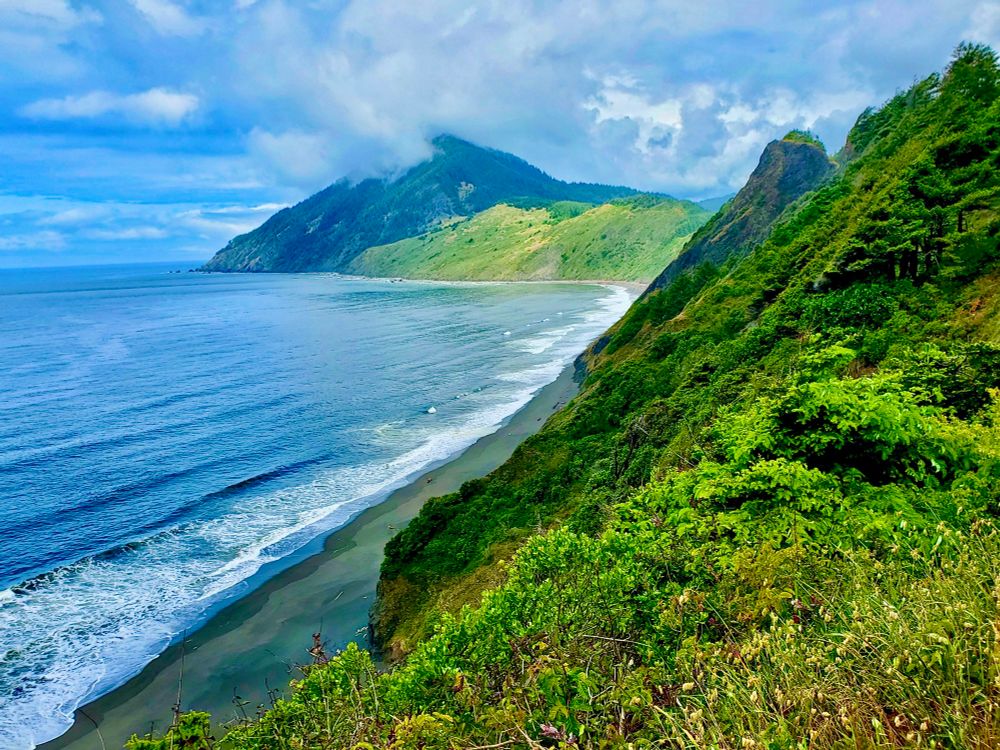 A mountain top view of the blue ocean with gentle white capped waves rolling in onto a dark sandy beach. The beach is at the bottom of green foliage covered mountains and curves around towards the left with a view of another mountain in the background. Fluffy white and blue clouds fill the sky overhead.
