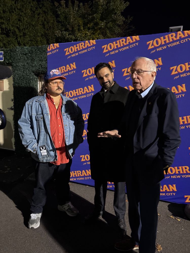 Photo credit Jeremy Slevin. 

Comedian Stavros Halkias, Mayoral Candidate and Democratic Nominee Assemblymember Zohran Mamdani, and United States Senator Bernie Sanders stand outdoors at night in front of a blue “Zohran for New York City” campaign backdrop. Halkias wears a denim jacket and cap, Mamdani wears a dark coat and smiles slightly, and Sanders, in glasses and a dark suit, gestures while speaking.
