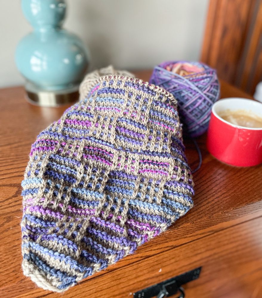 A knitting project in a grid of tan and purples sitting on a wooden table with a coffee cup and lamp in the background. 
