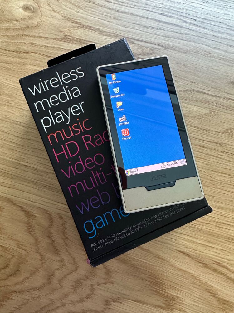 A silver Zune HD showing a Windows CE desktop, siting atop its box.